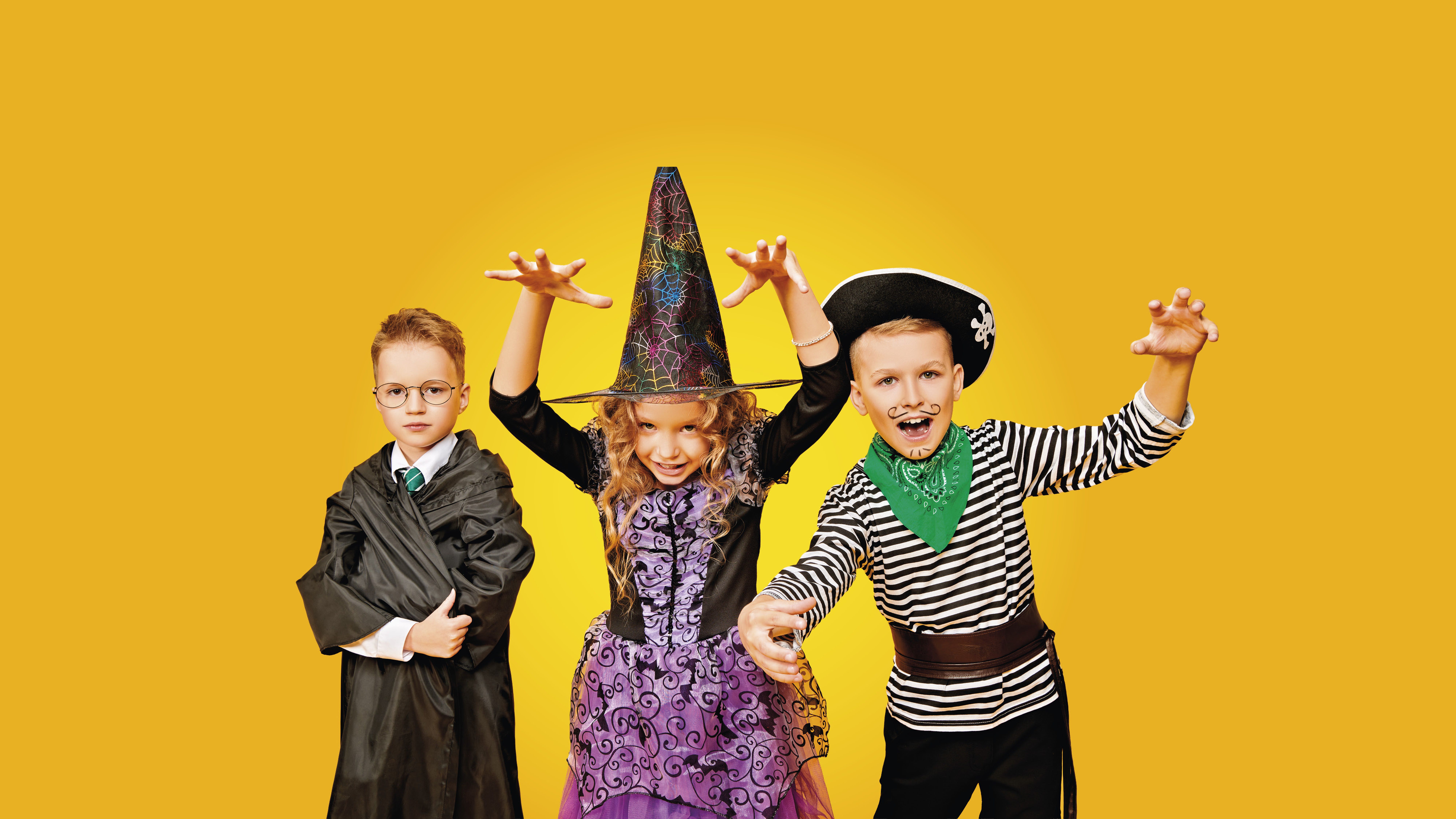 Na žlutém pozadí stojí tři děti v kostýmech: jedno v čarodějnickém rouchu, jedno v čarodějnickém oblečení a jedno v kostýmu piráta.