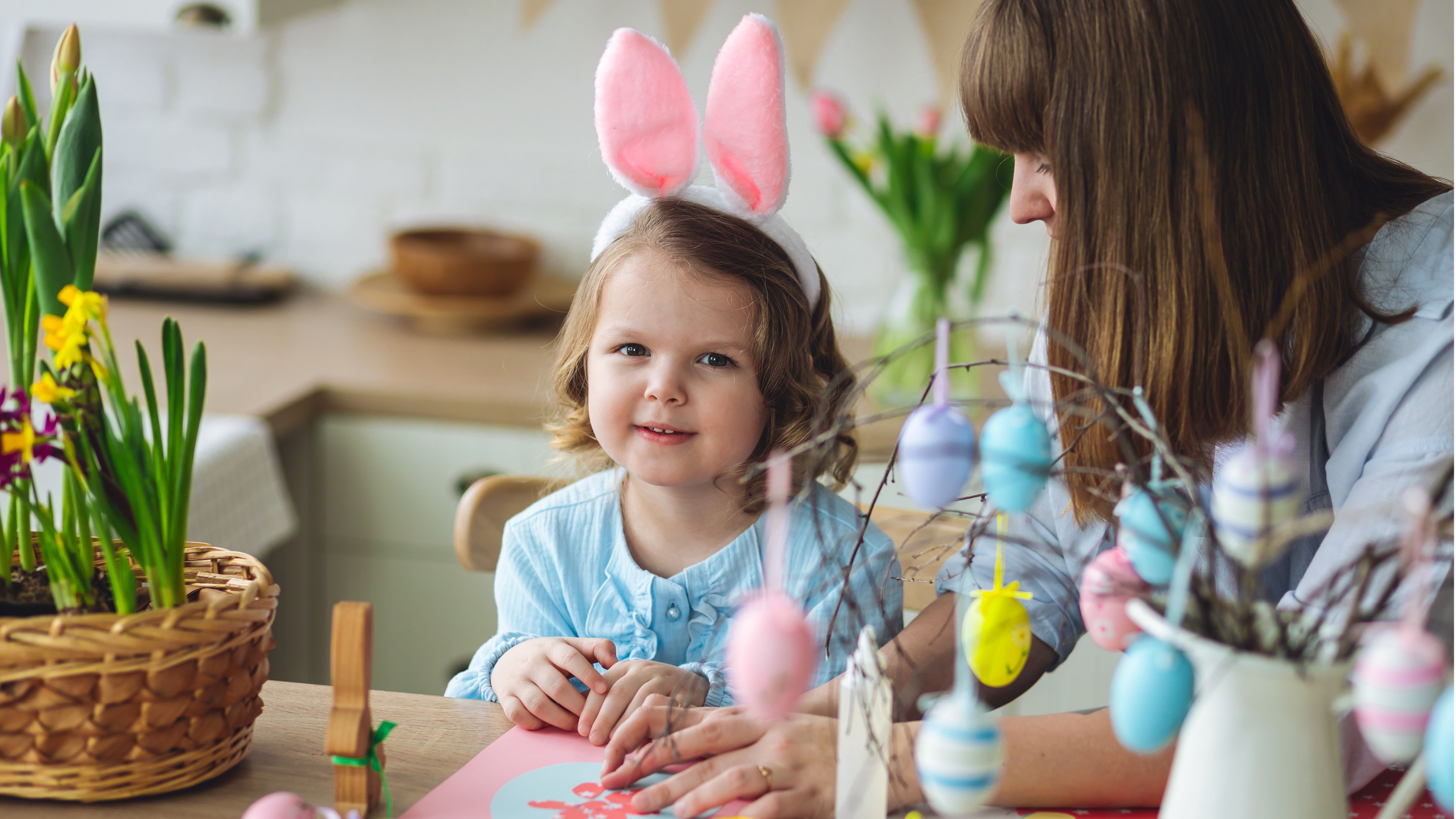 Dítě s růžovýma ušima a modrou košilí sedí u stolu vedle dospělé ženy. Barevná vajíčka zdobí větve v bílé váze.
