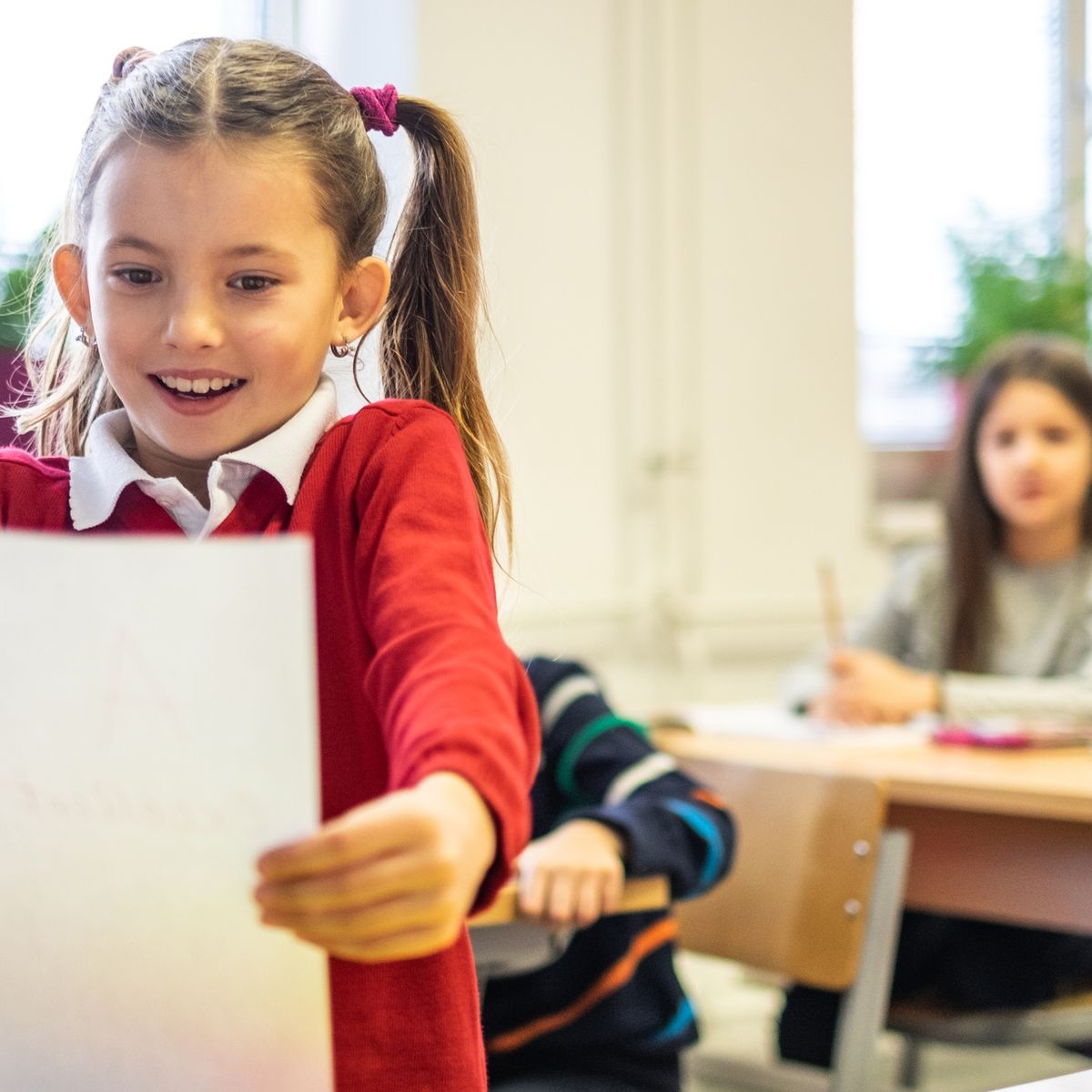 Usměvavá dívka v červeném svetru zvedá papír ve třídě a v pozadí pracují další dvě děti.