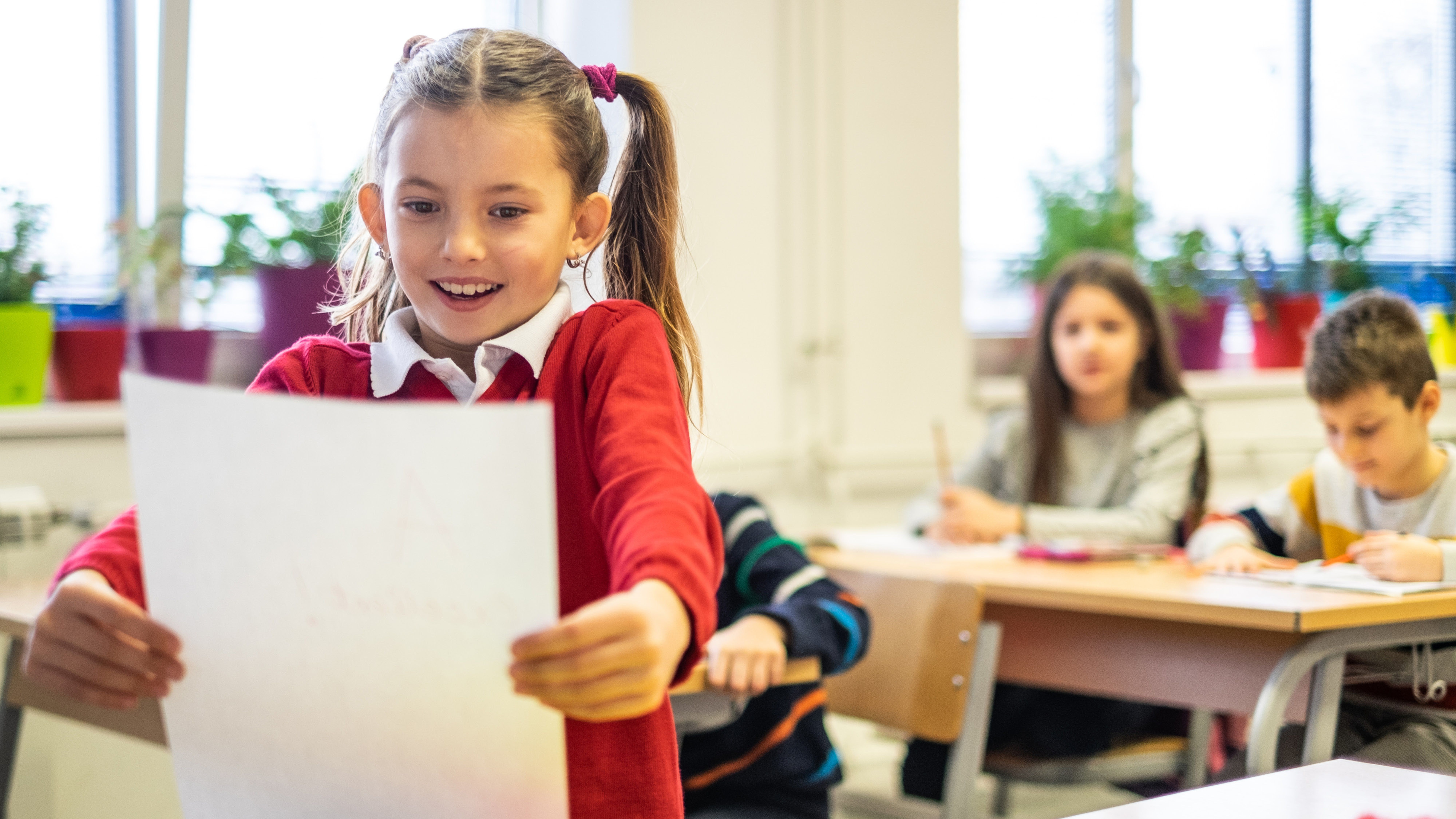 Usměvavá dívka v červeném svetru zvedá papír ve třídě a v pozadí pracují další dvě děti.