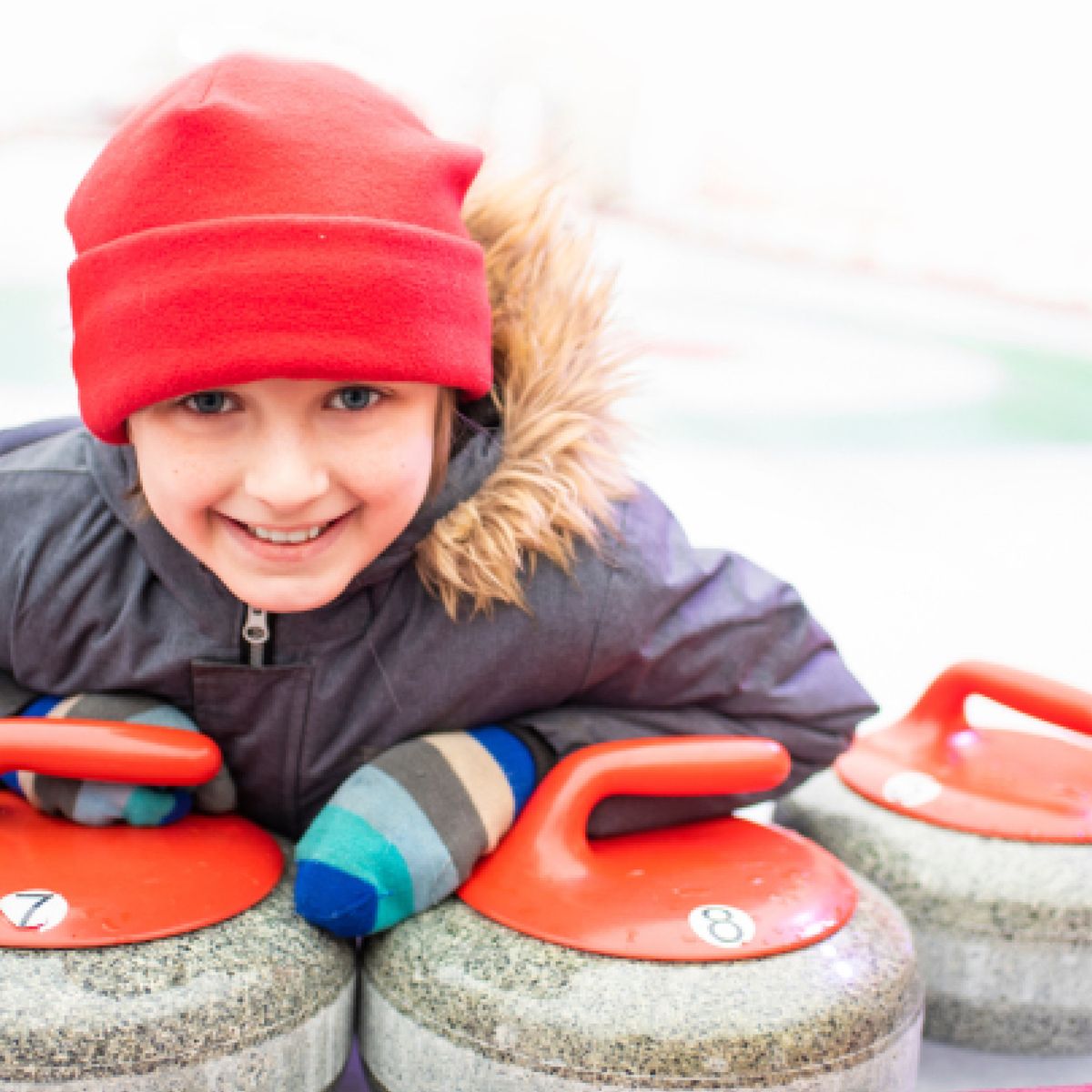 Dítě v červené čepici a modrých pruhovaných rukavicích se usmívá a opírá se o curlingové kameny s červenými držadly na kluzišti.