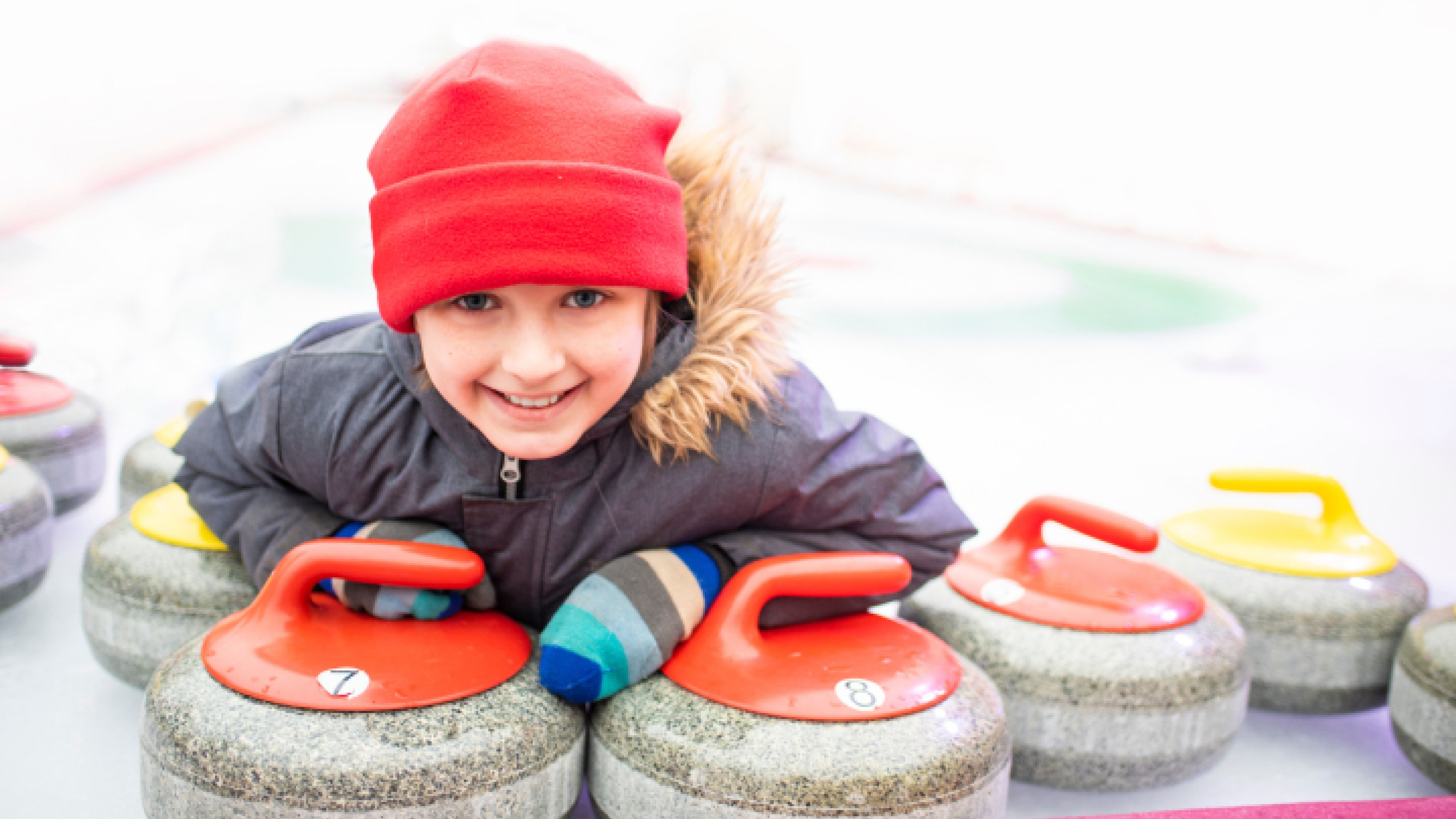 Dítě v červené čepici a modrých pruhovaných rukavicích se usmívá a opírá se o curlingové kameny s červenými držadly na kluzišti.