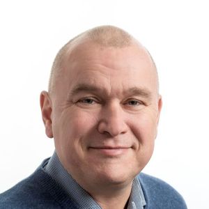 Osoba s vyholenou hlavou, oblečená v modrém svetru a kostkované košili, se usmívá na jednobarevném bílém pozadí.