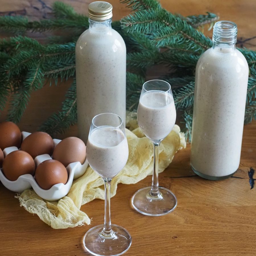 Dvě láhve a sklenice vaječného likéru na dřevěném stole s vejci, borovicovými větvičkami a žlutým ubrusem.