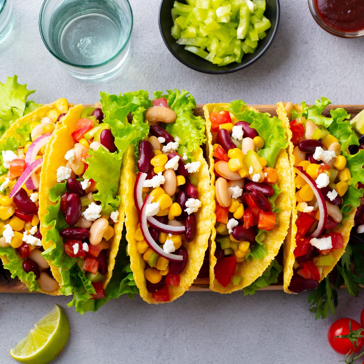 Čtyři barevné vegetariánské tacos s fazolemi, kukuřicí, salátem a sýrem na dřevěném prkénku, obklopené nakrájenými rajčaty, kousky limetky a různými přísadami.