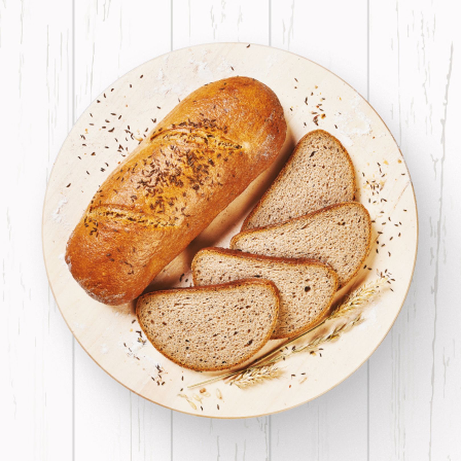 Bochník kmínového chleba a tři plátky na ozdobném talíři, položené na bílém dřevěném povrchu, najdete ho v Pekárně Globus..
