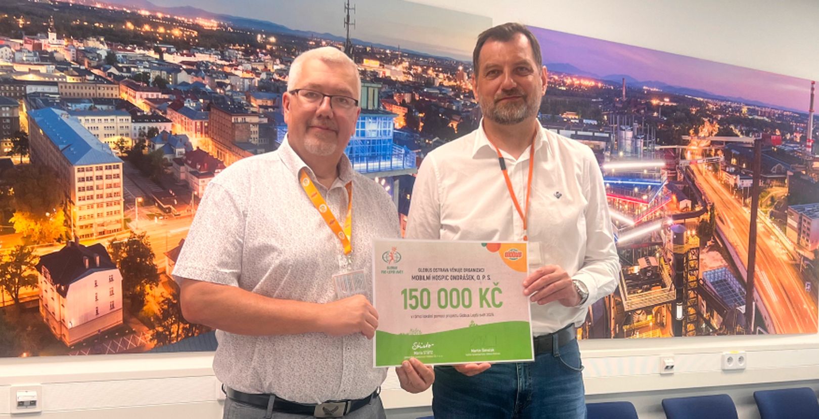 2 muži držící šek na 150 000 Kč před fotografií města