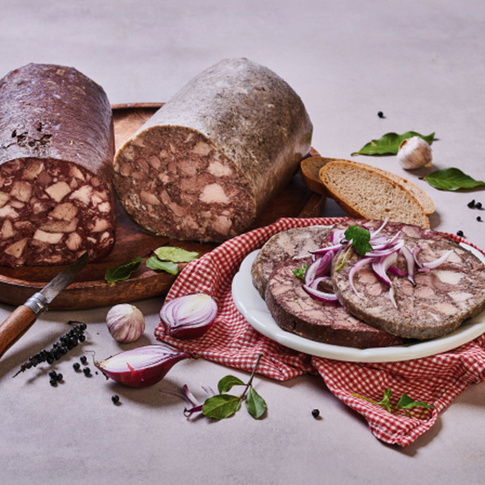 Dvě tlačenky (světlá a tmavá) na dřevěném prkénku, nakrájené kusy na talíři s cibulí, česnekem, kuličkami pepře a chlebem na červené utěrce.
