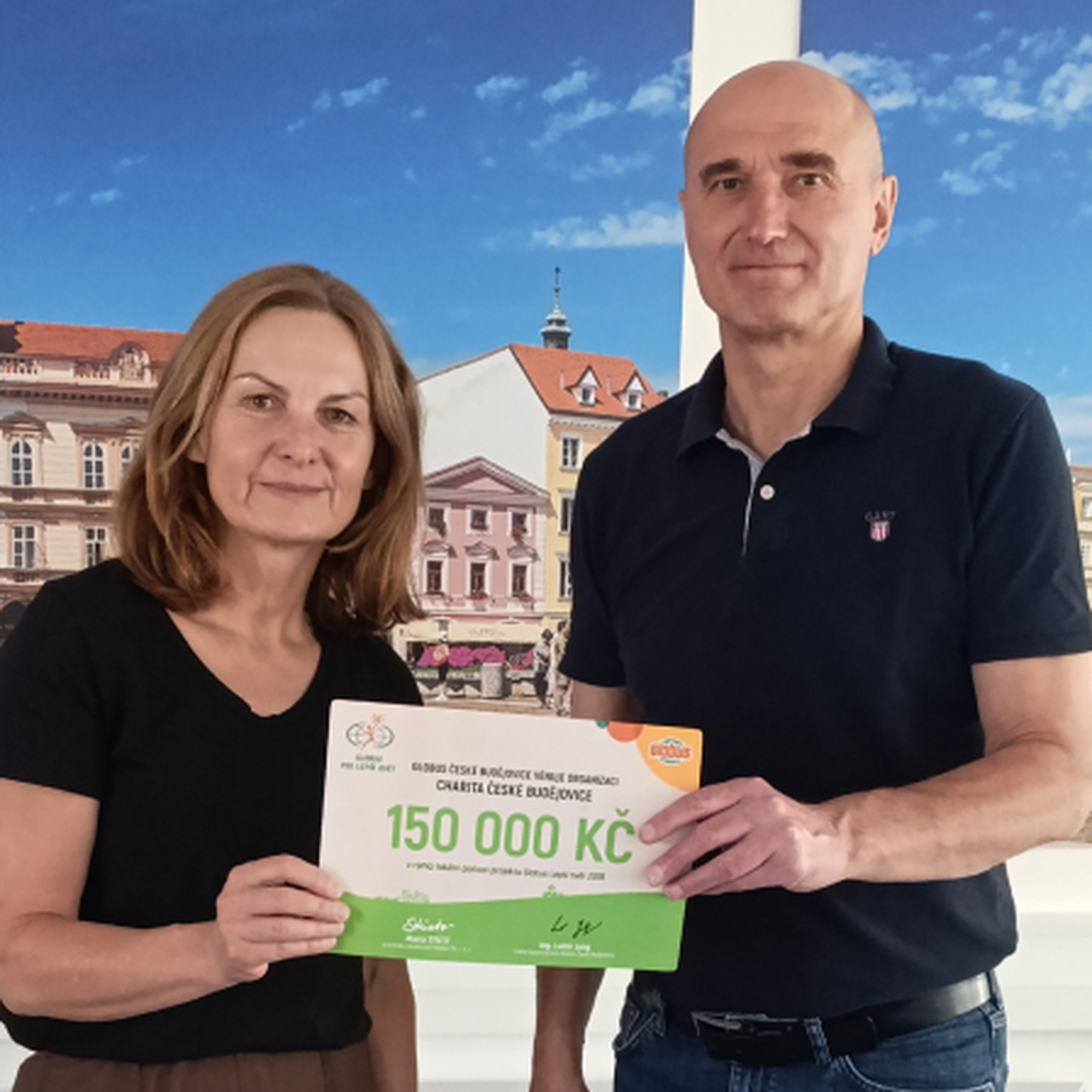 Muž a žena  s darovacím šekem na částku 150 000 Kč před fotkami budov