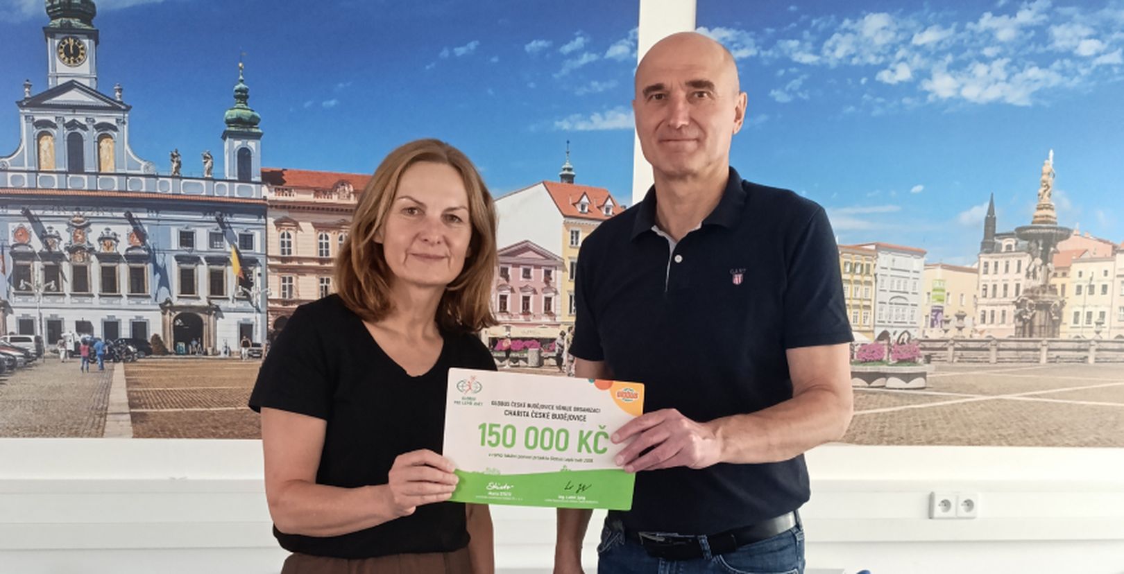 Muž a žena  s darovacím šekem na částku 150 000 Kč před fotkami budov