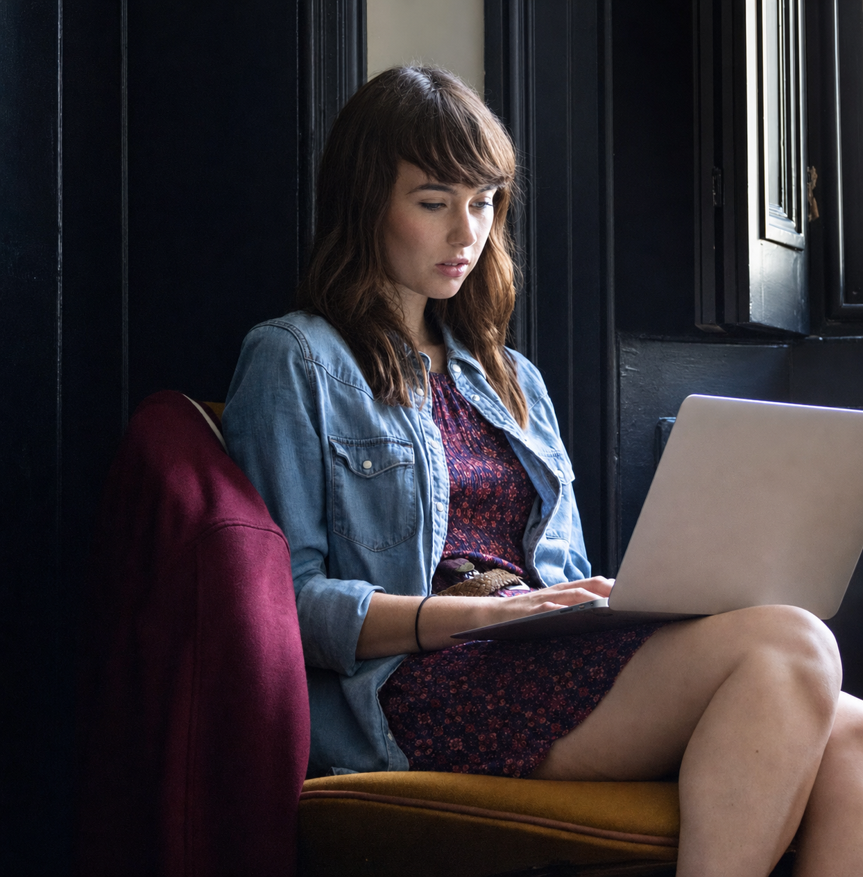 Woman sitting indoors using a laptop, wearing a denim jacket and floral dress, sunlight streaming through a window.