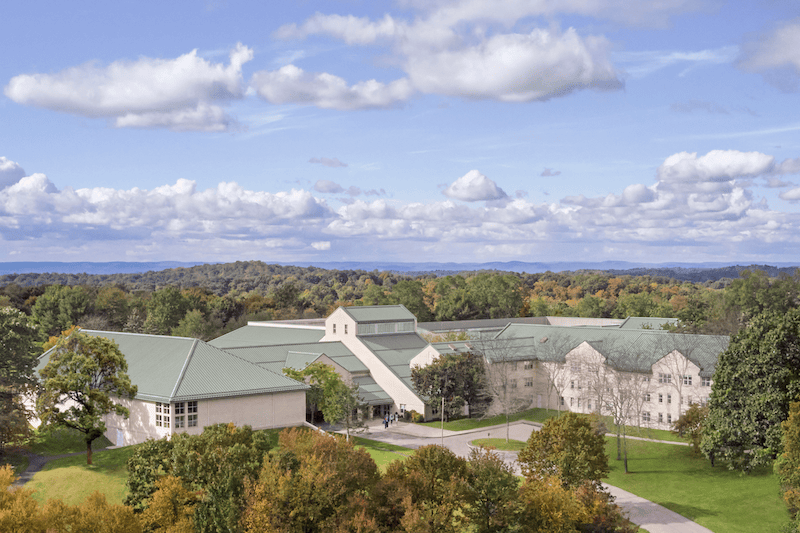 Aerial view of a large building complex surrounded by lush trees, with a clear blue sky and scattered clouds in the background.