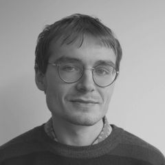 Young man with glasses and short hair, wearing a patterned shirt and a striped sweater, smiling softly against a plain background. Black and white photo.