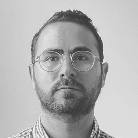 Black and white portrait of a man with glasses, a short beard, and plaid shirt, looking directly at the camera against a plain background.