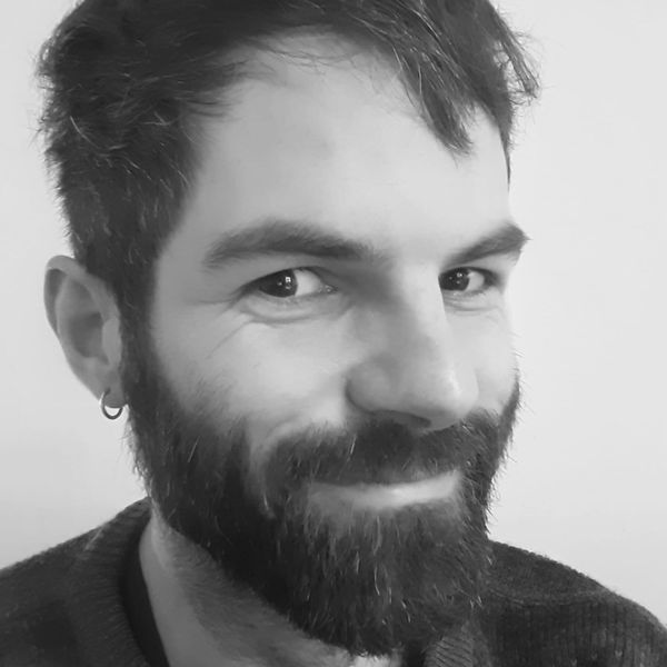 Man with a beard and earring, smiling slightly, looking towards the camera against a plain background.