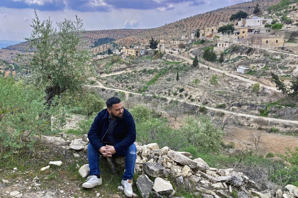 Man sitting on a rock, overlooking a rural hillside village with scattered houses and winding dirt paths under a cloudy sky.