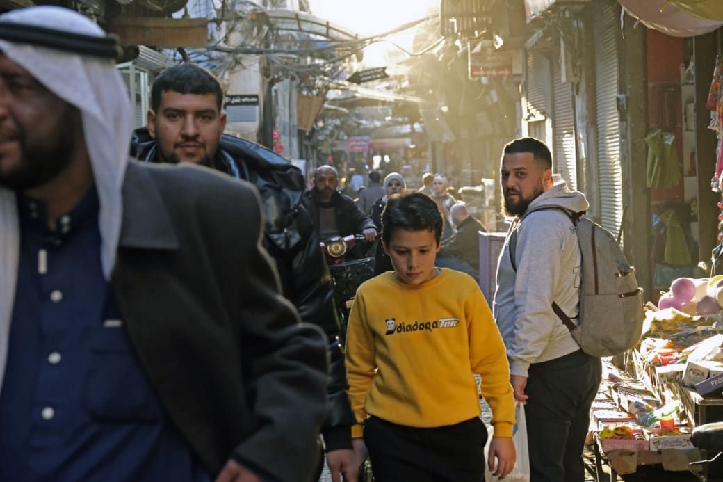 Busy street scene with people walking through a narrow alley. Sunlight filters through overhead, highlighting the bustling atmosphere.