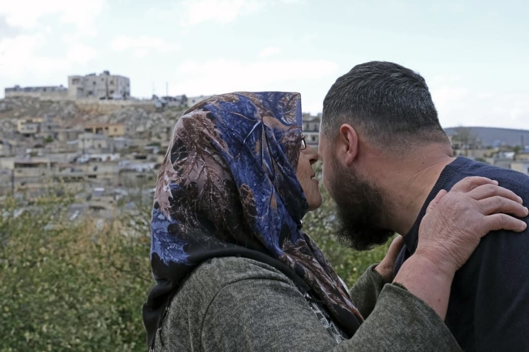 A bearded man and a woman in a headscarf embrace closely, with a village and hillside in the background under a partly cloudy sky.