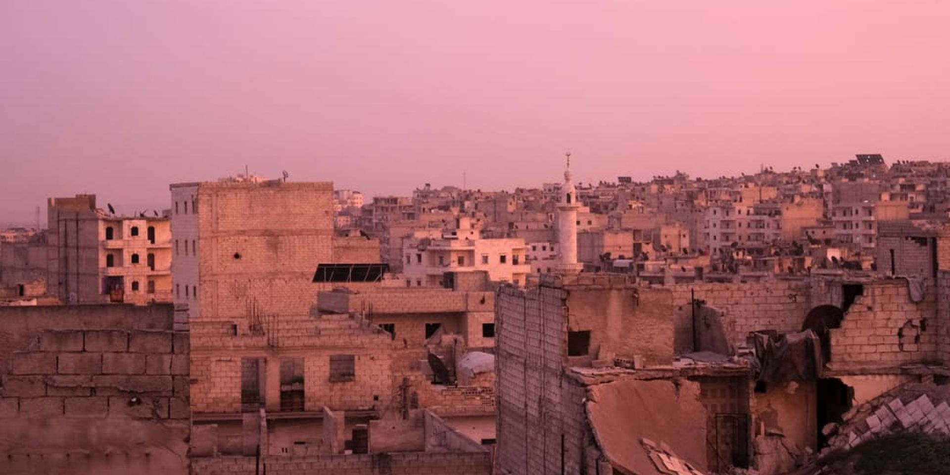 Urban landscape at sunset with damaged buildings under a pink sky, featuring a prominent minaret in the background.