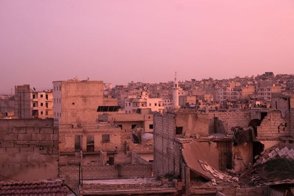 Urban landscape at sunset with damaged buildings under a pink sky, featuring a prominent minaret in the background.