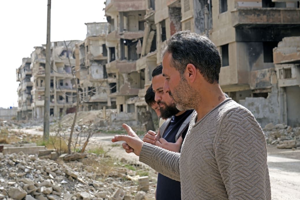 Two men walking amidst ruins of partially destroyed buildings, pointing towards the debris-filled street in a war-torn area.