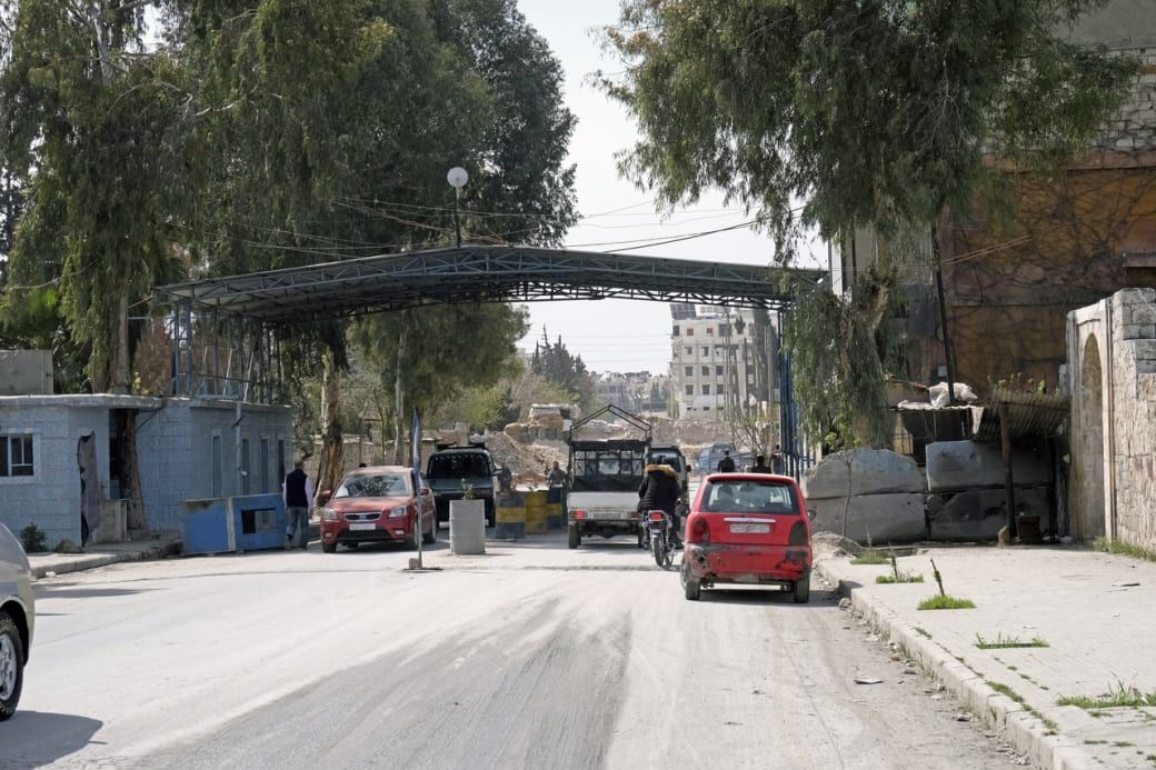 A busy checkpoint with vehicles and motorbikes passing through, surrounded by trees and buildings under a metal canopy.