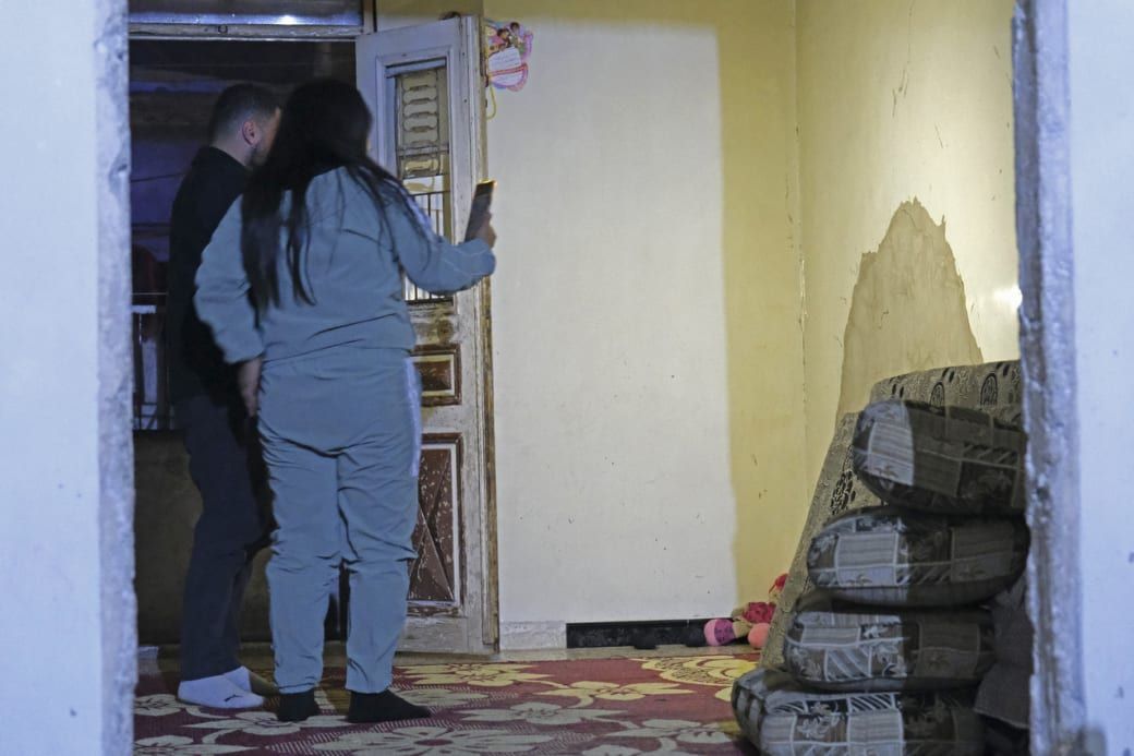 Two people stand near a partially open door in a dimly lit room with stacked cushions and peeling wall paint.