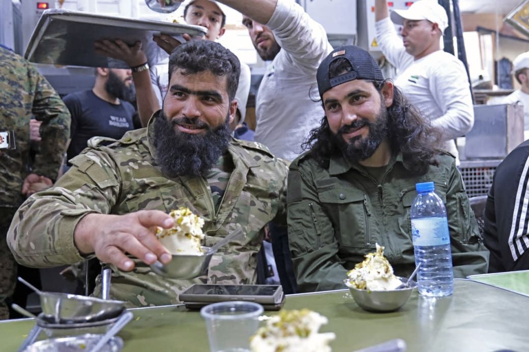 Two men in military attire enjoy a meal with dessert, surrounded by others in a busy setting.