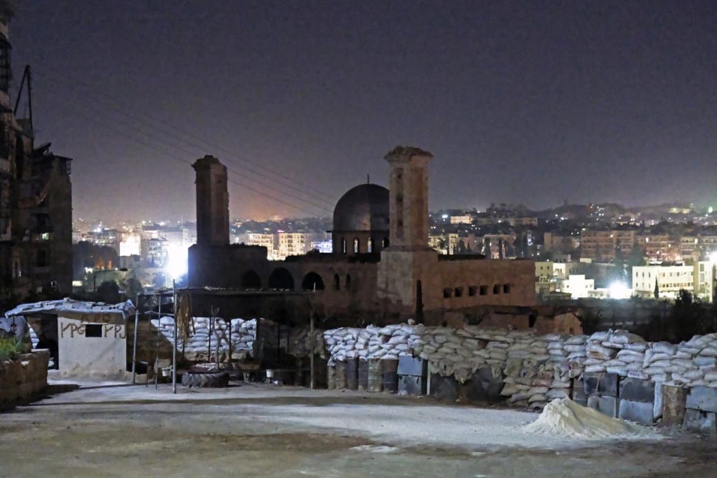 Night view of a partially ruined building with two towers and a dome, surrounded by sandbags, overlooking a lit-up cityscape.