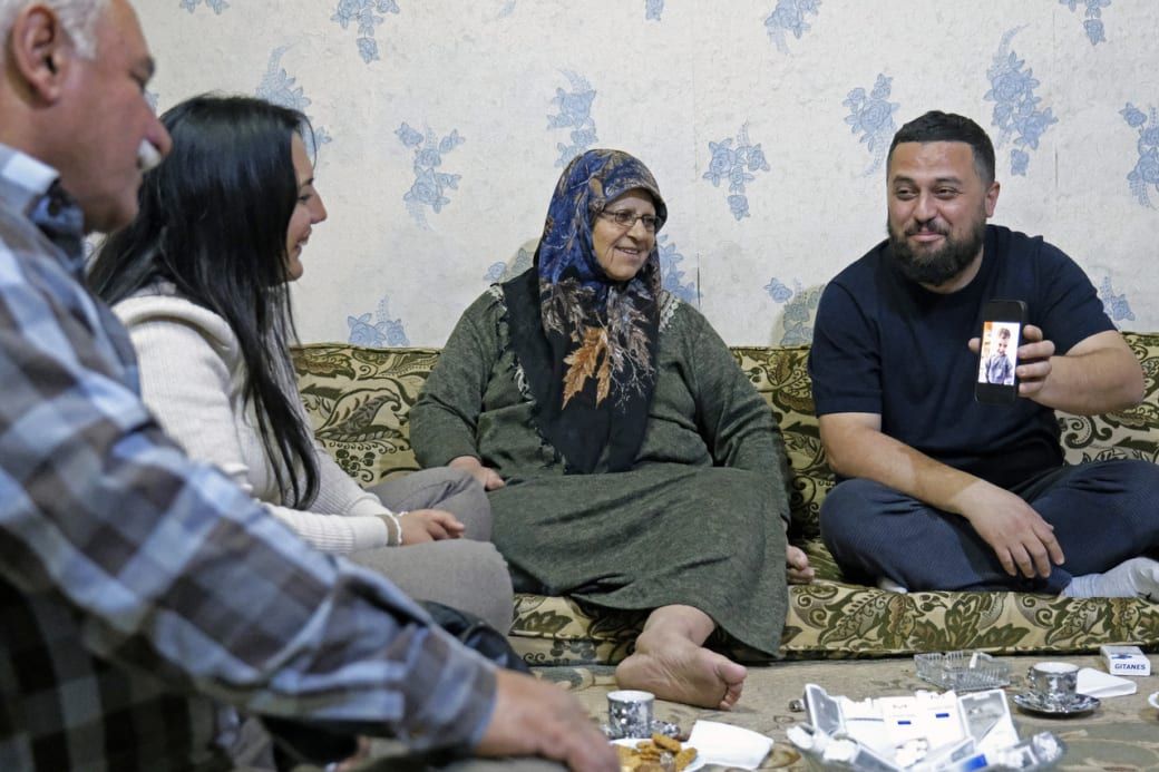 A family gathers on a patterned couch, smiling as a man shows a photo on his phone, with snacks and tea on the table.