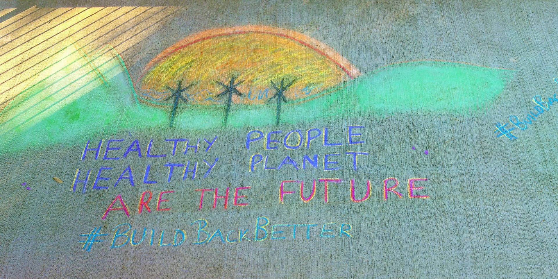 Chalk drawing with a sun, trees, and text saying, "Healthy people, healthy planet are the future. #BuildBackBetter."