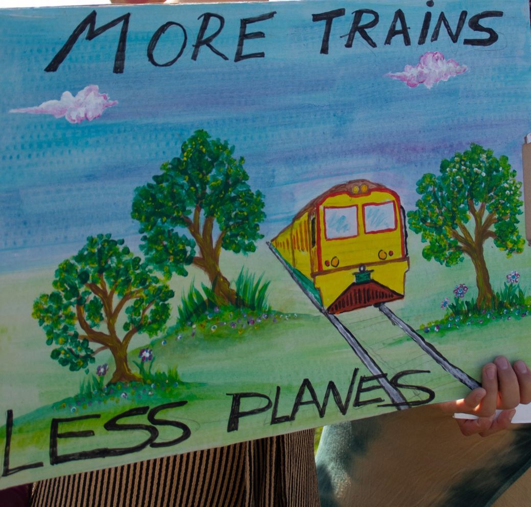 Hand-painted sign depicting a train with trees, reading "More Trains Less Planes" against a blue sky with clouds.