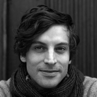 Smiling man with wavy hair, wearing a dark scarf, and a light sweater, set against a dark, textured background.