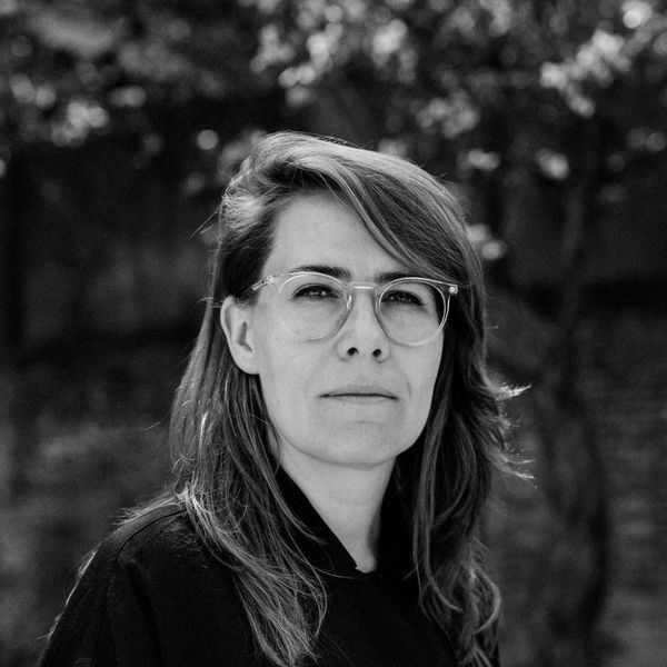 Black and white portrait of a person with glasses, long hair, and a serious expression, standing outdoors with blurred trees in the background.