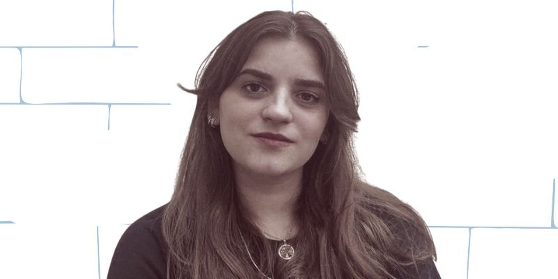 Woman with long hair wearing a dark shirt, standing against a white brick-patterned background.