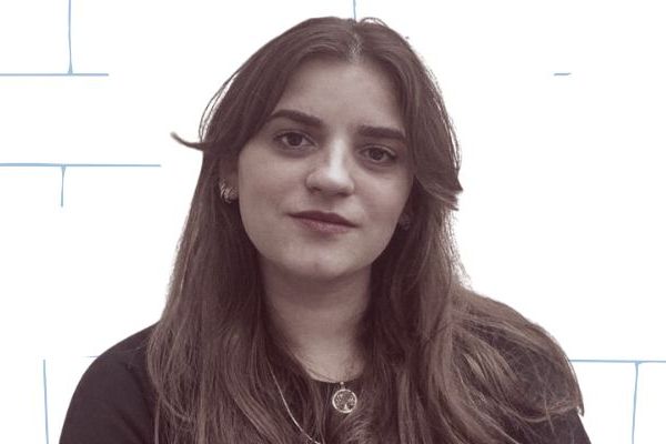 Woman with long hair wearing a dark shirt, standing against a white brick-patterned background.