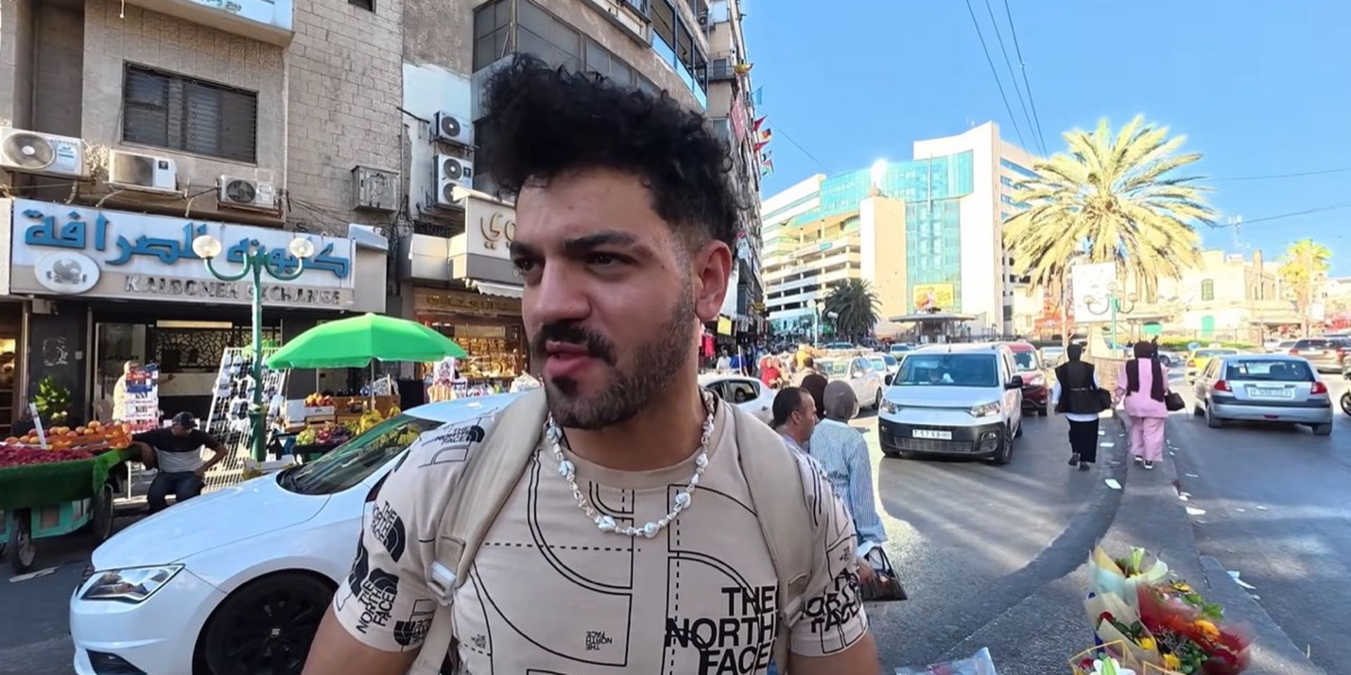 A man with dark hair and a patterned shirt stands on a busy urban street with cars, buildings, and market stalls in the background.