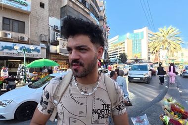 A man with dark hair and a patterned shirt stands on a busy urban street with cars, buildings, and market stalls in the background.