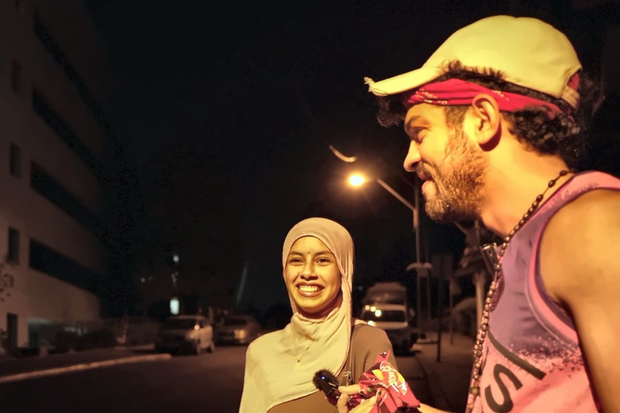 Two people, a woman in a hijab and a man with a cap, smile and talk on a dimly lit street at night.