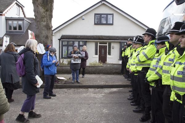 A line of police officers faces a group of protesters holding signs on a residential street. A house and tree are visible in the background.