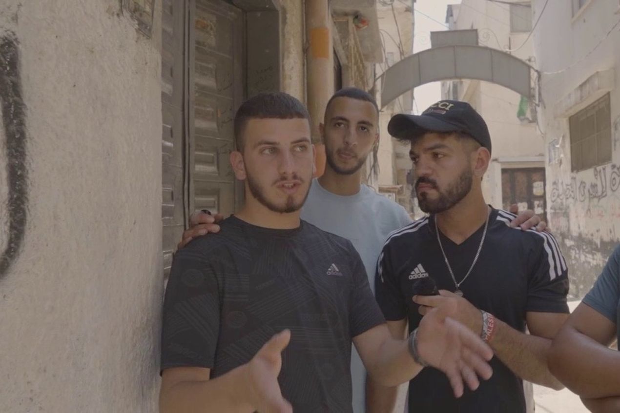 Four young men stand in a narrow street, one gesturing with his hands while talking, with the others listening attentively.
