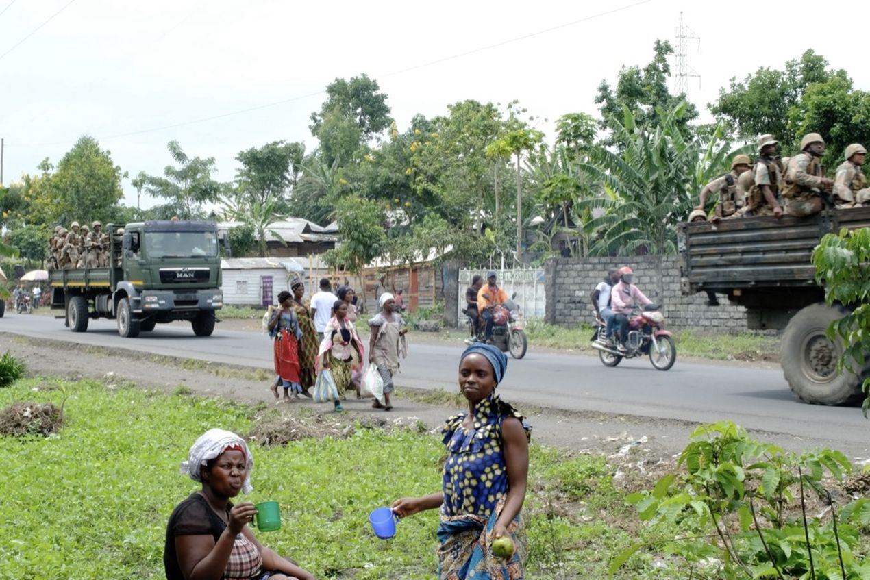 Women stand by the roadside while military trucks and motorcycles pass, set against a backdrop of greenery and houses.