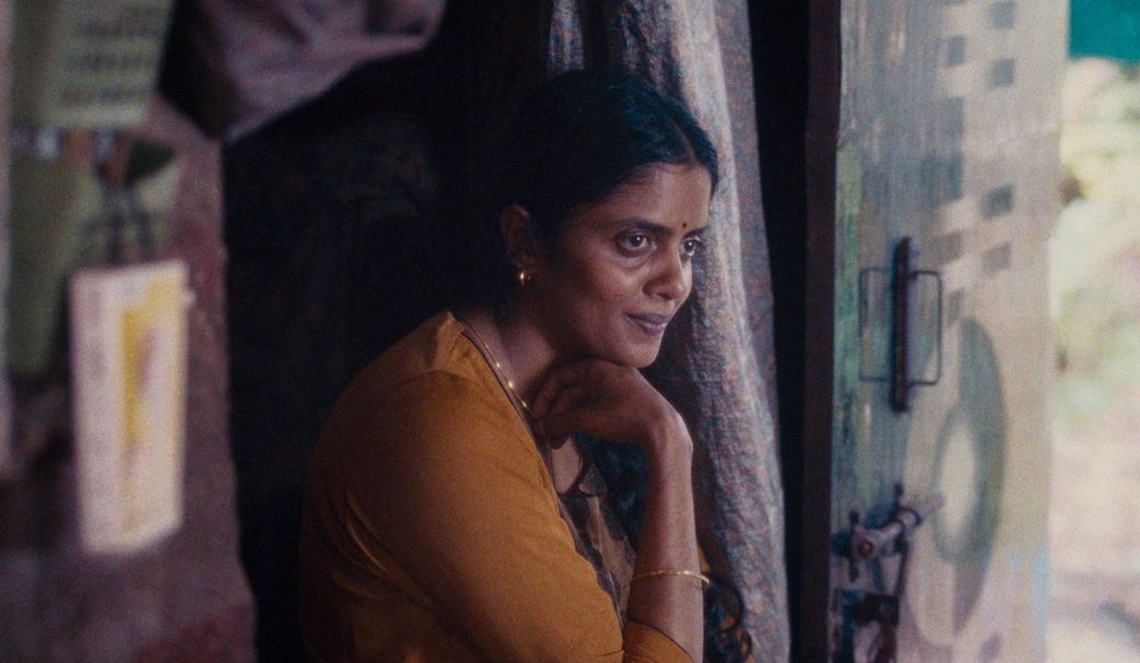 A woman in a yellow outfit and gold jewelry sits pensively by a doorway, resting her chin on her hand, with a thoughtful expression.