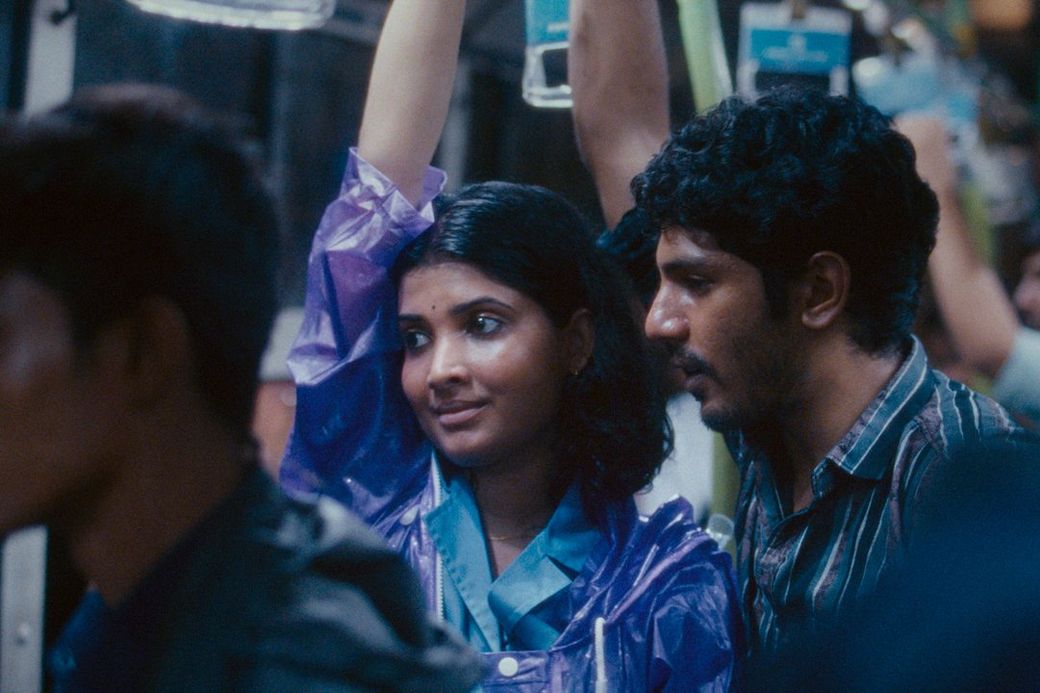 A woman in a purple raincoat holds an overhead strap inside a crowded bus, standing close to a man in a striped shirt.