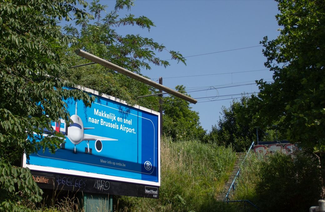 A billboard near greenery advertises easy travel to Brussels Airport. It features an image of an airplane and text in Dutch.