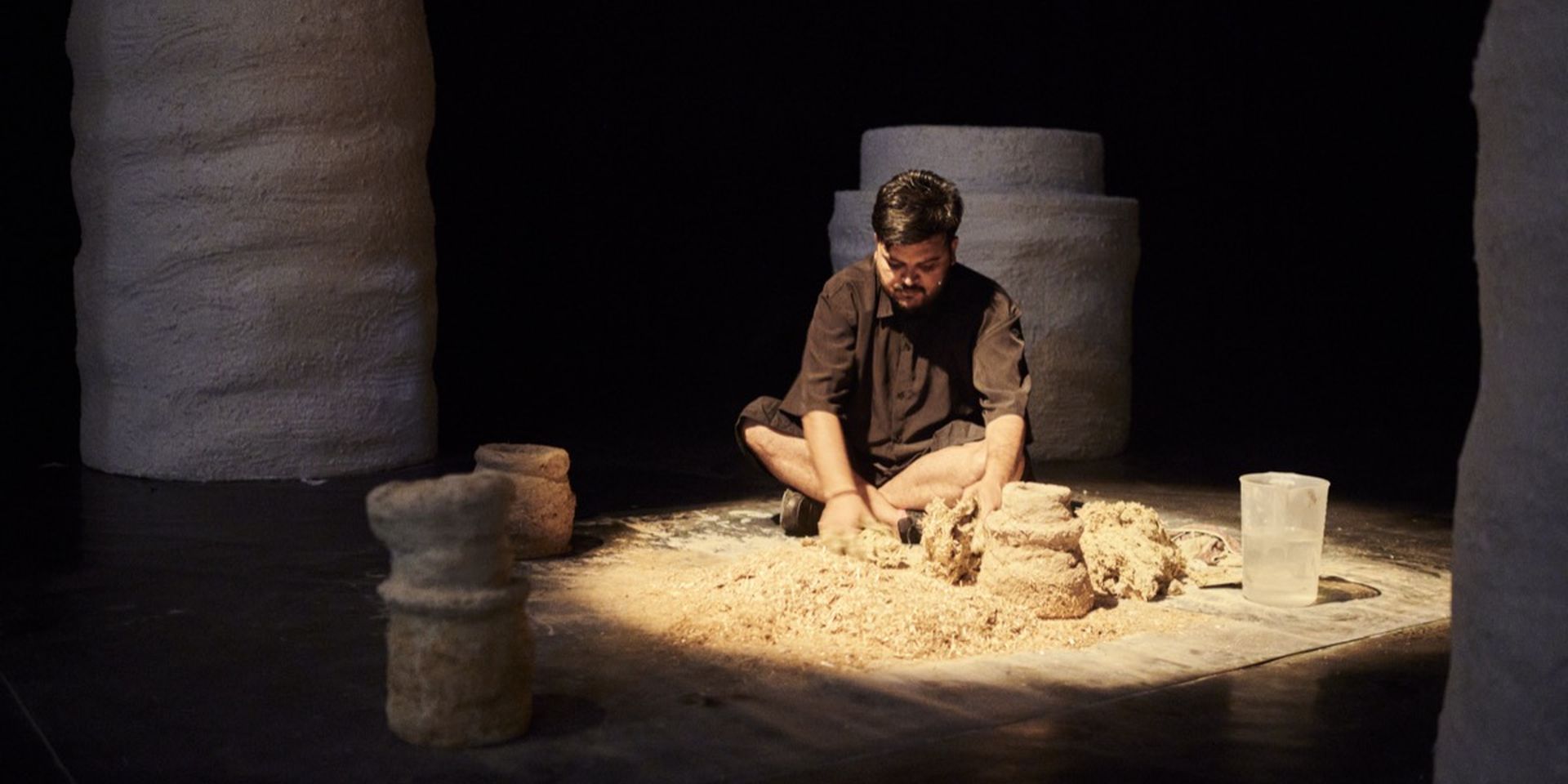 A man in black clothing kneels on the floor, shaping clay surrounded by large, textured pillars in a dimly lit space.