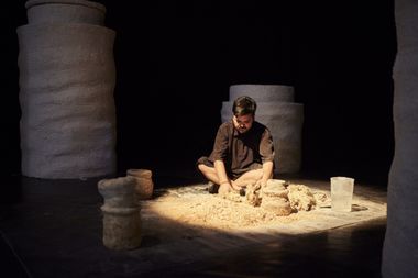 A man in black clothing kneels on the floor, shaping clay surrounded by large, textured pillars in a dimly lit space.