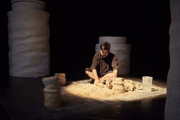A man in black clothing kneels on the floor, shaping clay surrounded by large, textured pillars in a dimly lit space.