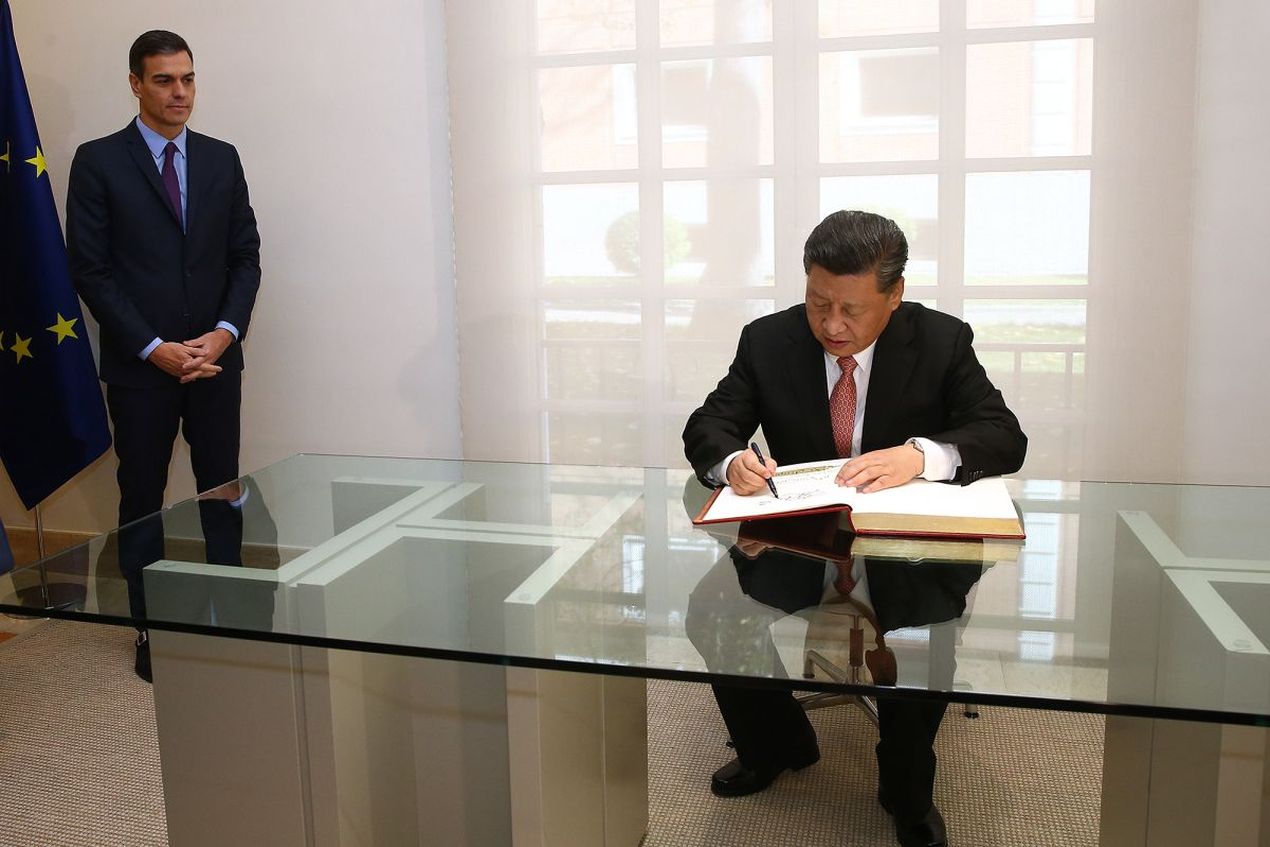 Spaans premier Pedro Sánchez staat links, tegen de muur aan, met gevouwen handen. Chinees president Xi Jinping zit, centraal in beeld, aan een bureau en schrijft iets in een dik boek. Foto uit 2018.