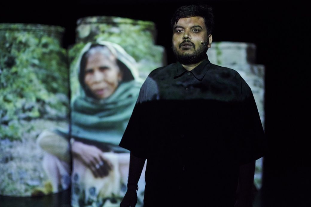 A person stands in dim lighting with an image of an elderly woman in a green shawl projected on cylindrical screens behind them.